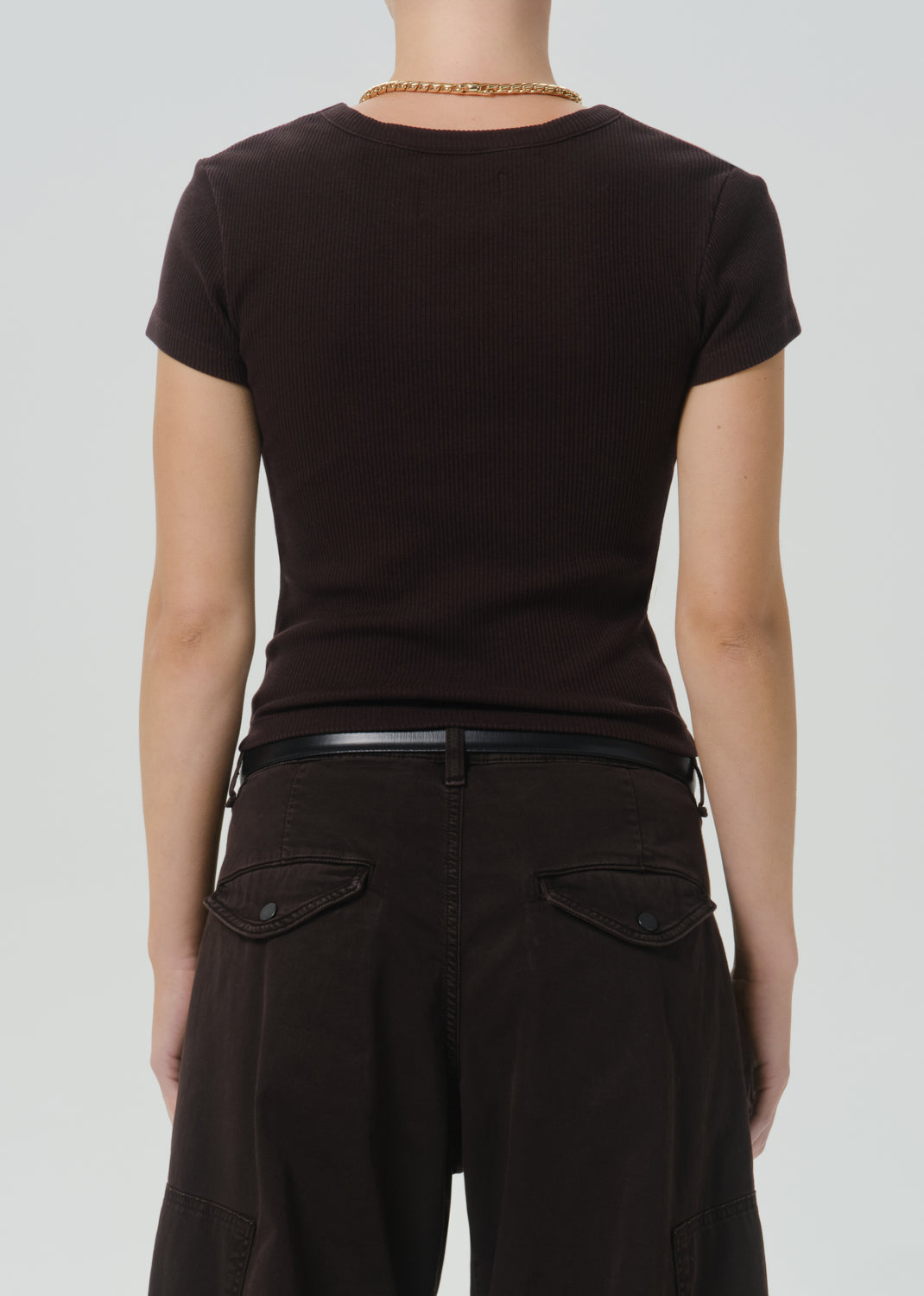 Rear view of a person wearing a fitted short-sleeve dark brown t-shirt and dark brown pants with a thin black belt, against a grey background.