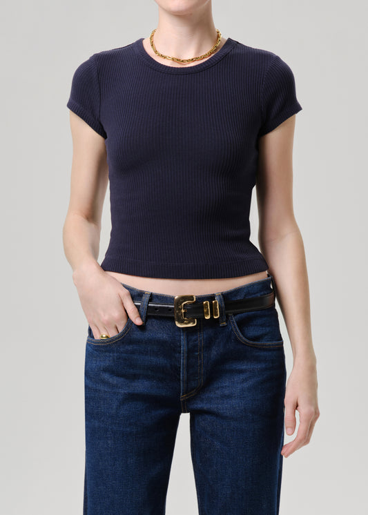 A model wearing a dark navy ribbed short-sleeve crop top paired with dark blue jeans and a black belt with a gold buckle, against a grey background.
