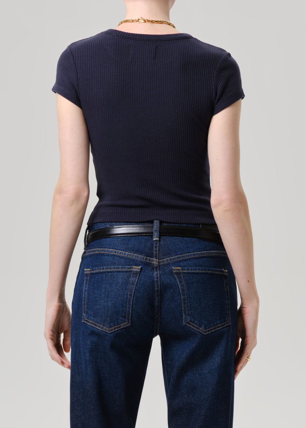 Rear view of a model wearing a dark navy ribbed short-sleeve top and dark blue jeans with a black belt, against a grey background.