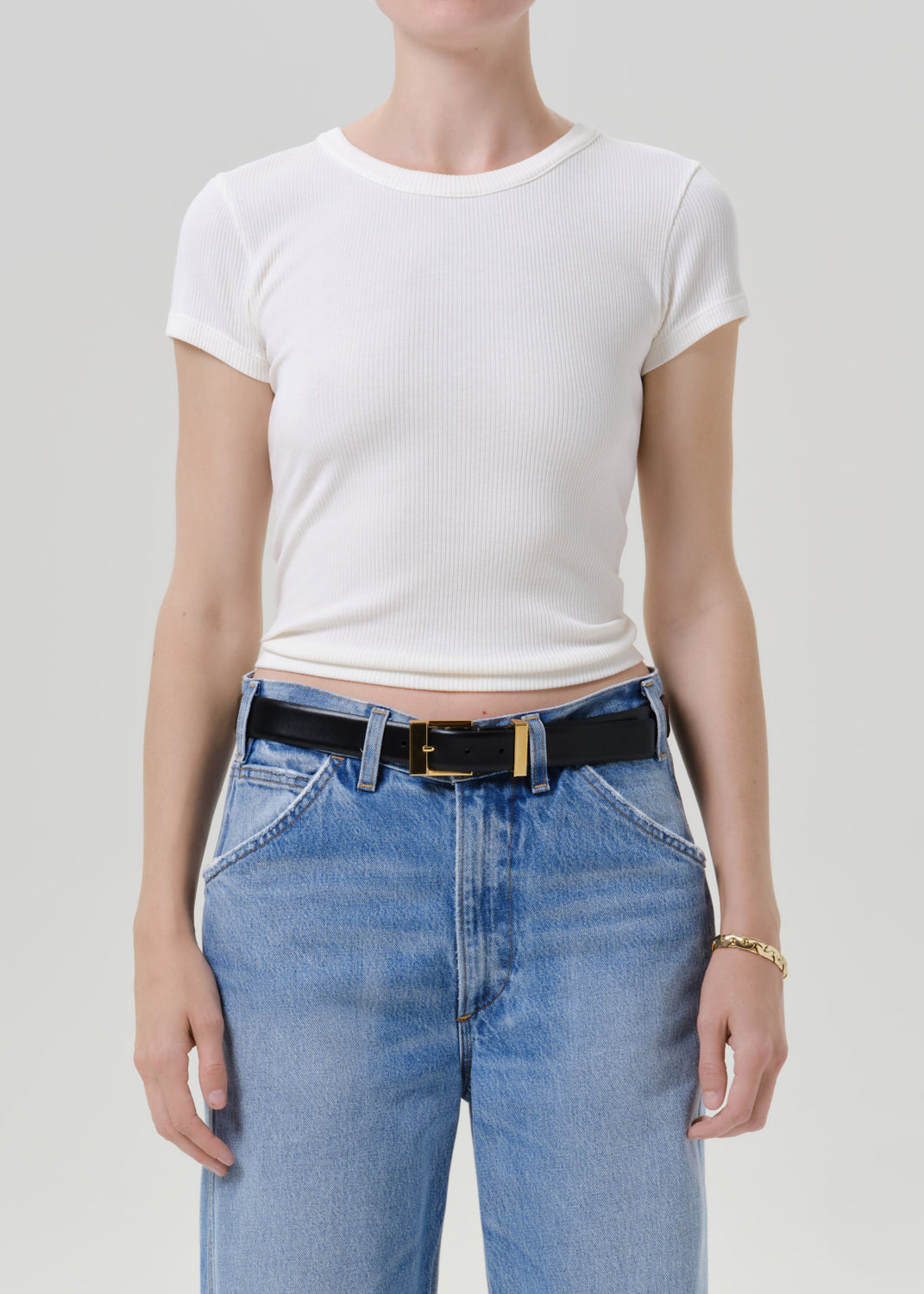 A model wearing a white short-sleeve ribbed crop top and light blue jeans with a black belt, shown from the neck down against a grey background.