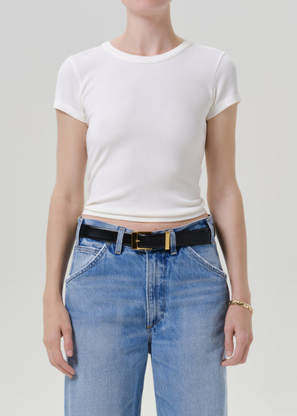 A model wearing a white short-sleeve ribbed crop top and light blue jeans with a black belt, shown from the neck down against a grey background.