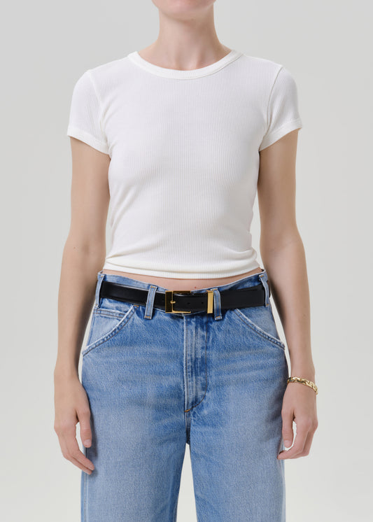A model wearing a white short-sleeve ribbed crop top and light blue jeans with a black belt, shown from the neck down against a grey background.