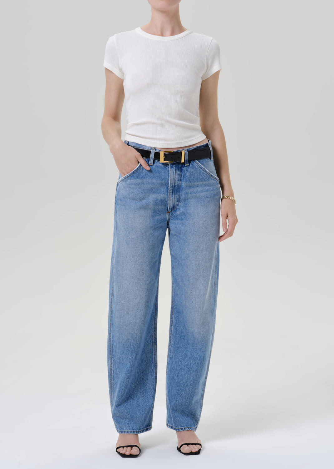 A model wearing a white short-sleeve crewneck t-shirt, light-wash blue wide-leg jeans with a black belt, and black sandals, standing against a grey background.