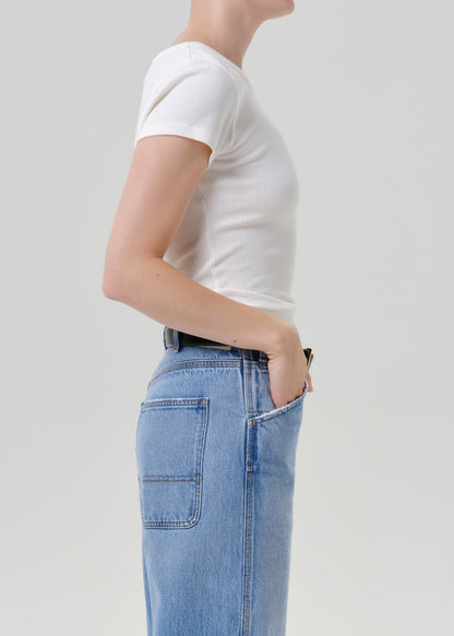 Side profile of a model wearing a white short-sleeve ribbed t-shirt tucked into light blue denim jeans with a black belt, against a grey background.