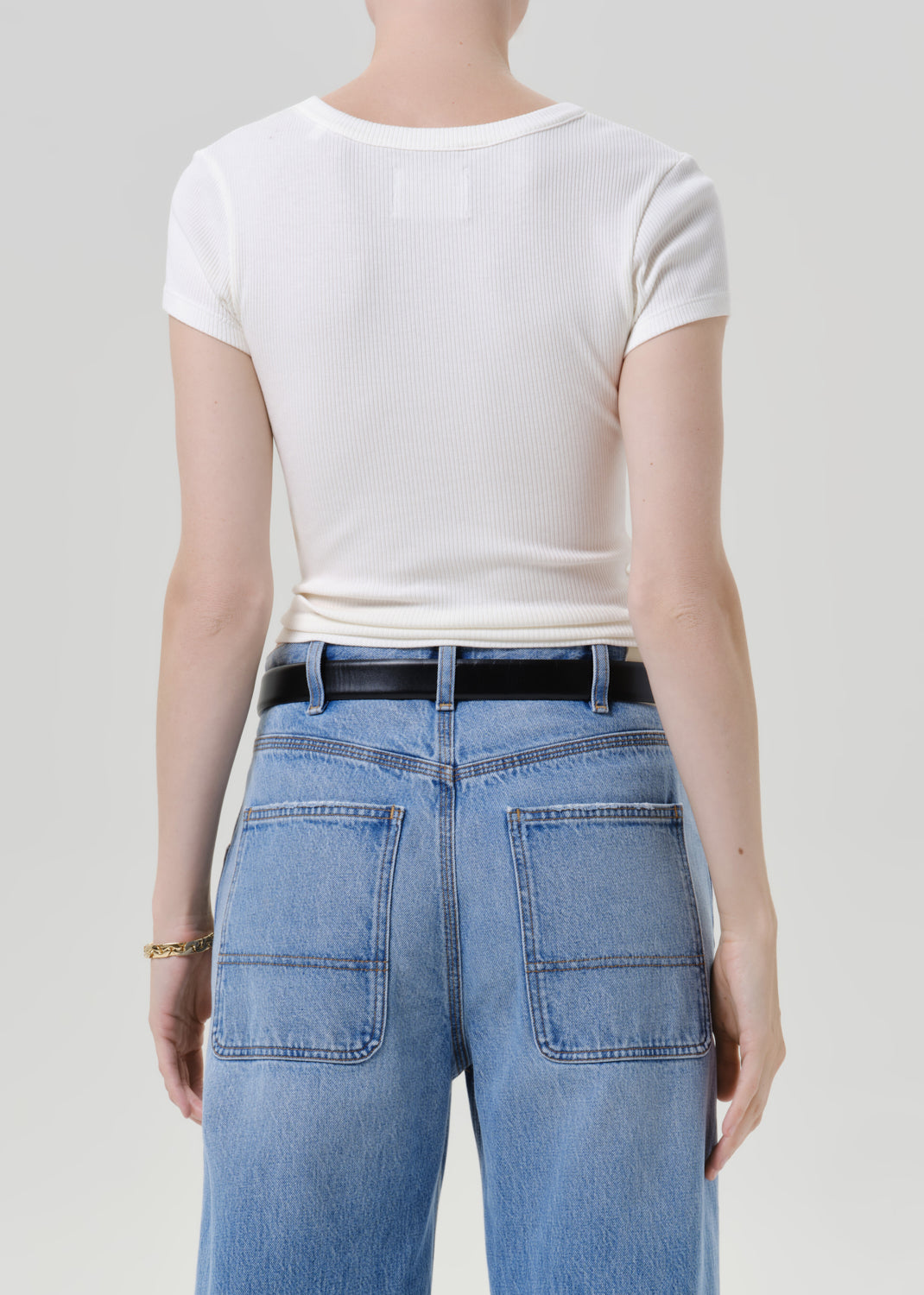 Rear view of a model wearing a white short-sleeve crewneck t-shirt tucked into light blue jeans with a black belt, against a grey background.