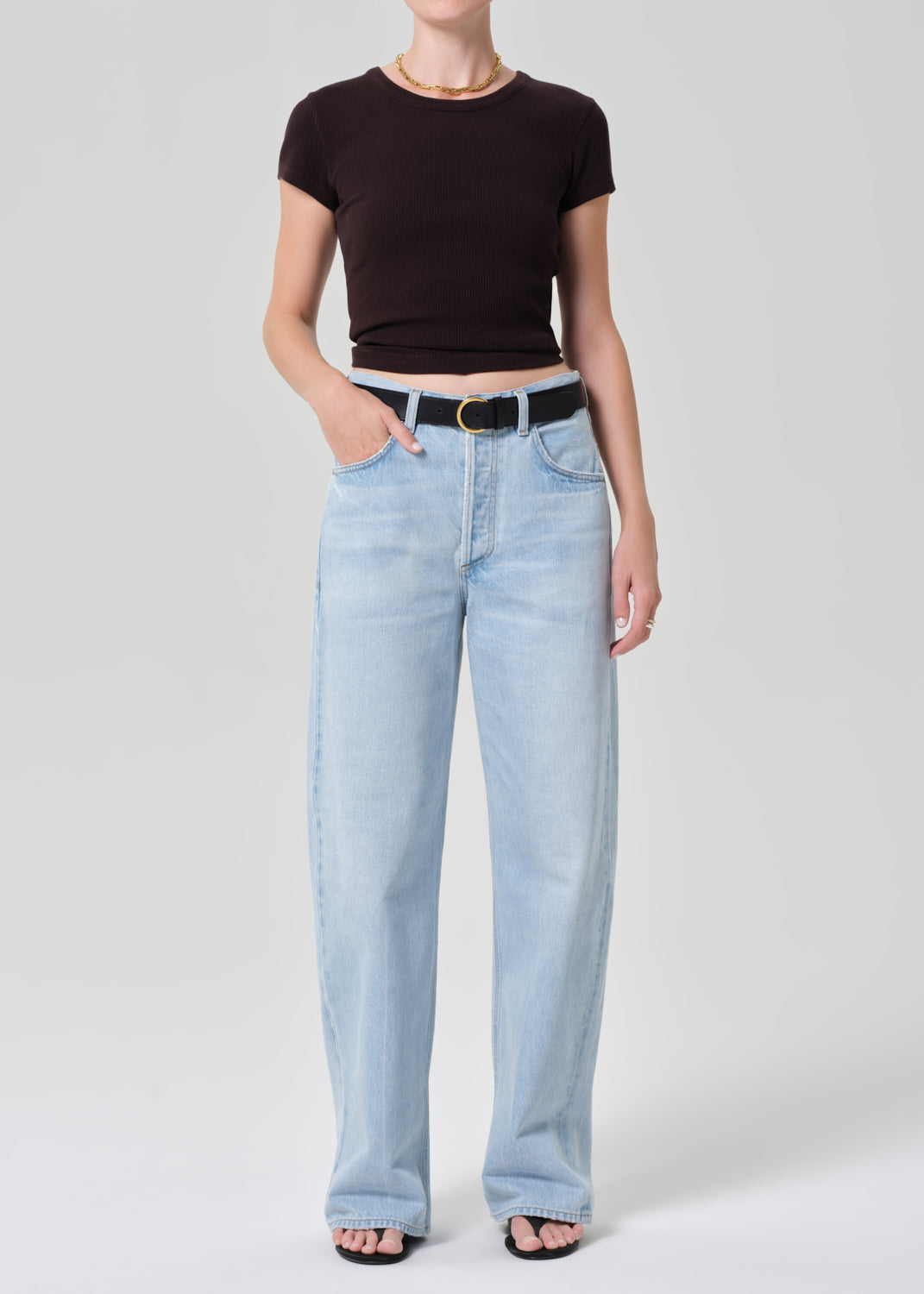 Person wearing a brown tee and light blue jeans with a black belt on a plain background