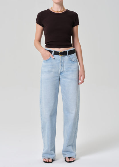 Person wearing a brown tee and light blue jeans with a black belt on a plain background