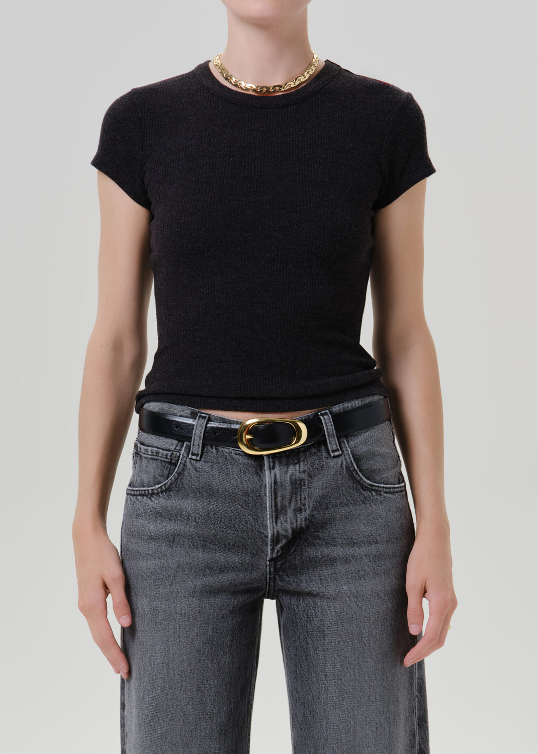 A model wearing a black short-sleeve crewneck knit top tucked into dark grey jeans with a black belt and gold buckle, accessorized with a chunky gold chain necklace, against a grey background.