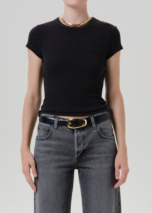 A model wearing a black short-sleeve crewneck knit top tucked into dark grey jeans with a black belt and gold buckle, accessorized with a chunky gold chain necklace, against a grey background.