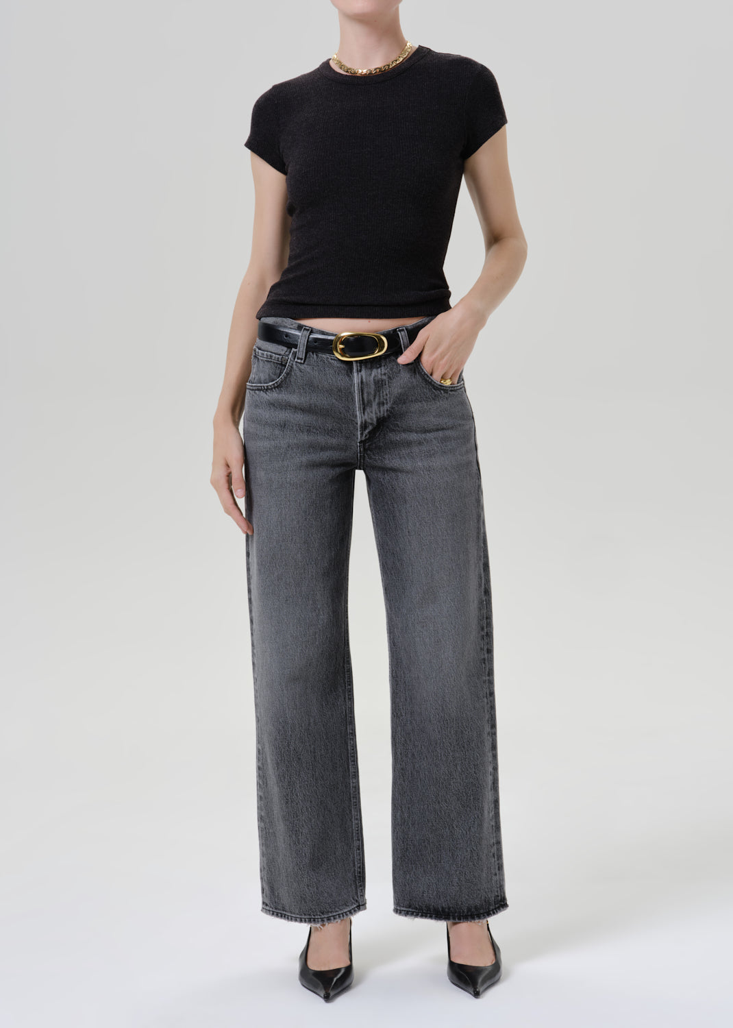A model wearing a black short-sleeve crewneck top, wide-leg grey denim jeans with a gold-tone belt, and black pointed-toe heels, photographed against a light grey background.