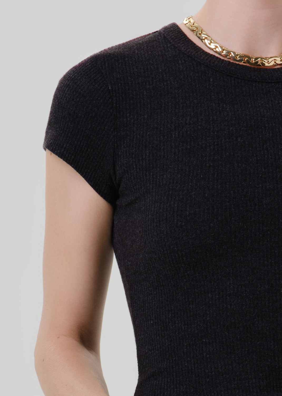 Close-up of a person wearing a dark charcoal ribbed short-sleeve top with a chunky gold chain necklace, against a grey background.