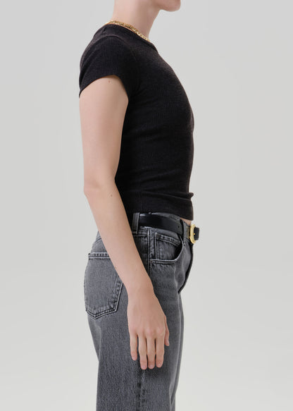 Side profile of a model wearing a black short-sleeve ribbed t-shirt tucked into grey jeans with a black belt and gold buckle, against a grey background.