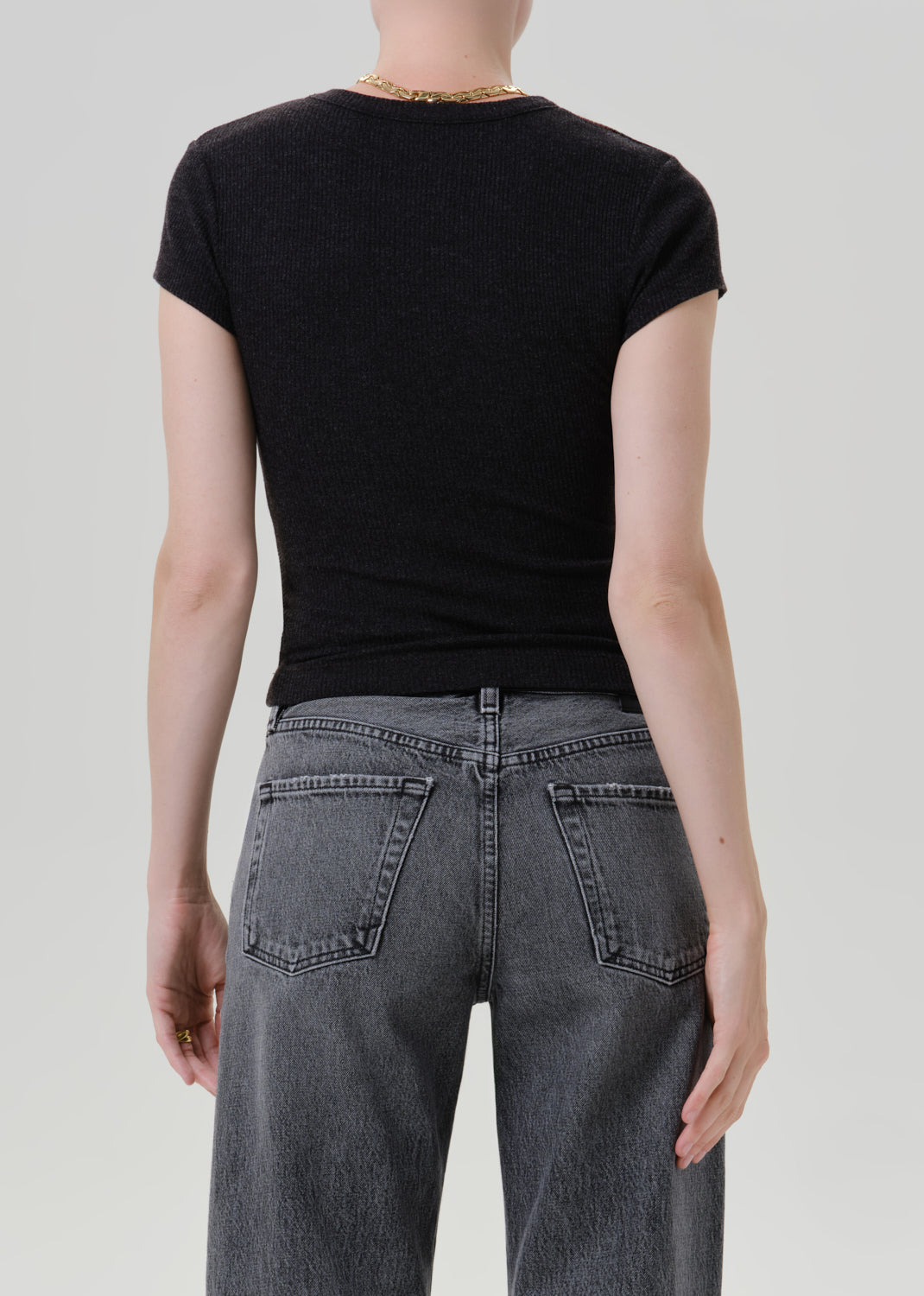Rear view of a person wearing a fitted black short-sleeve t-shirt and dark grey jeans against a light grey studio background.