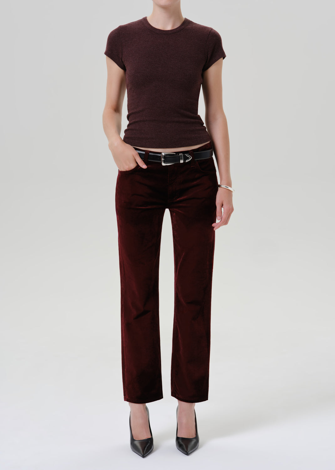 A model wearing a dark burgundy short-sleeve knit top and matching wide-leg trousers in a velvet-like fabric, styled with a black belt and black pointed-toe heels against a grey background.