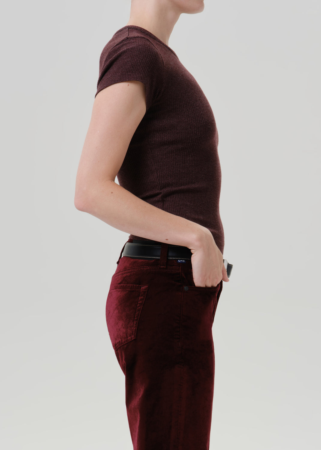 Side profile of a model wearing a short-sleeve ribbed knit t-shirt in dark burgundy and matching burgundy pants with a black belt, against a grey background.