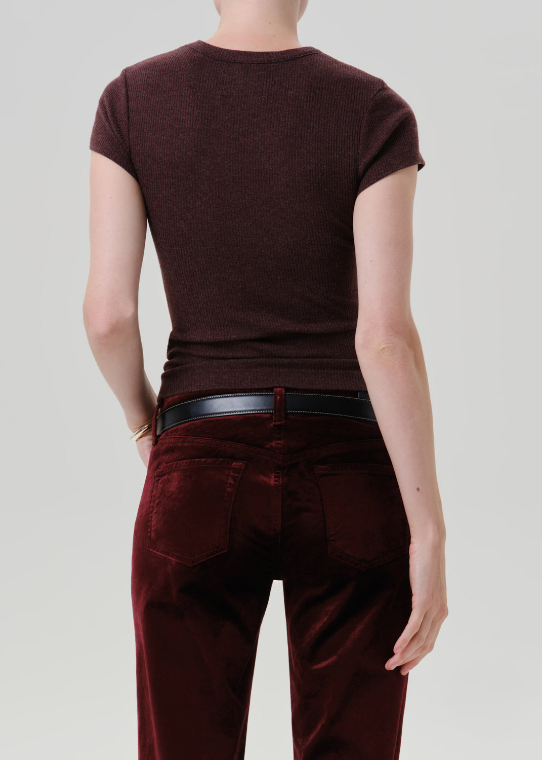 Rear view of a model wearing a dark burgundy ribbed short-sleeve t-shirt and matching burgundy velvet trousers, accessorized with a black leather belt, against a grey background.