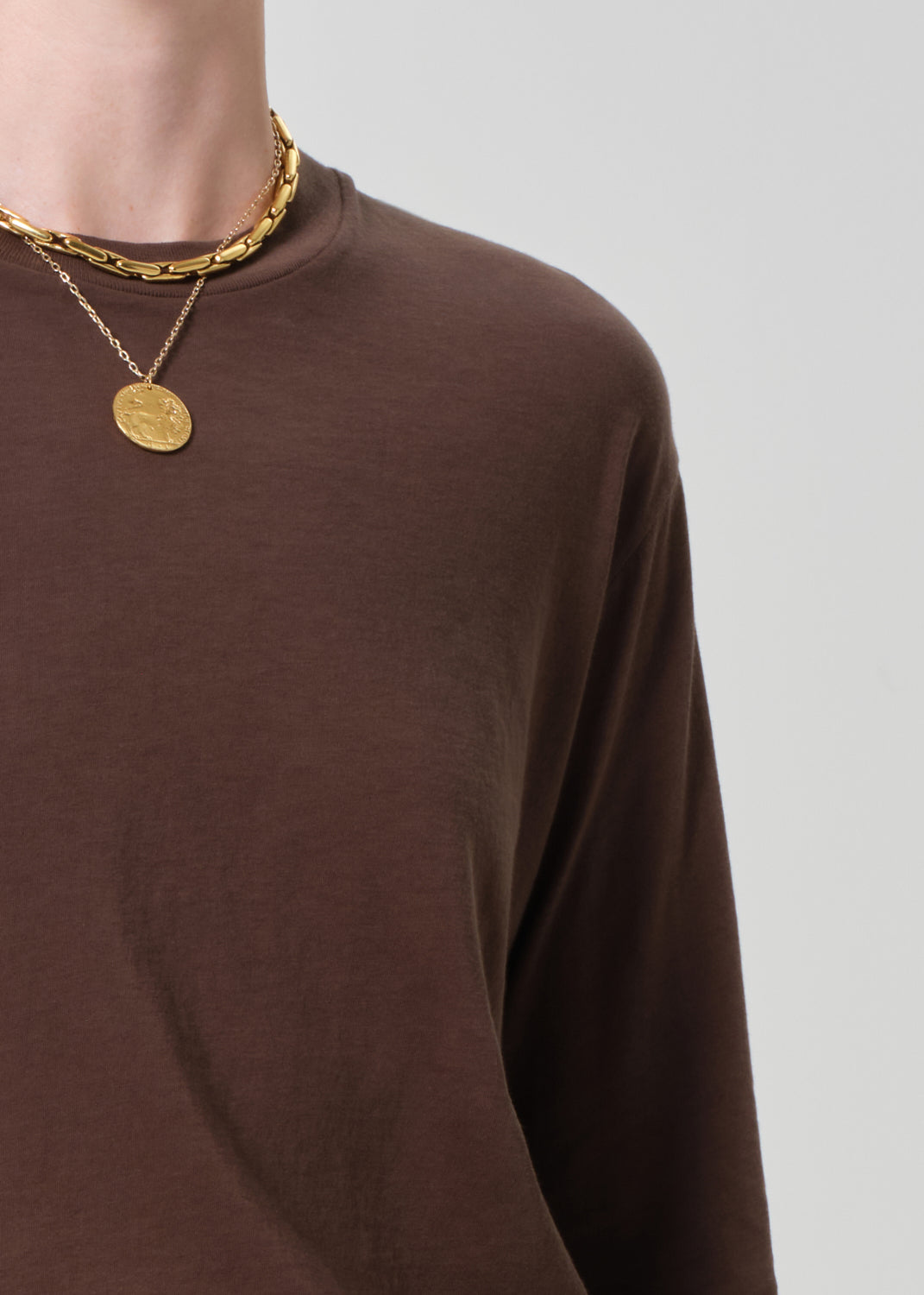 Person wearing a brown long-sleeve shirt with gold necklaces on a light gray background