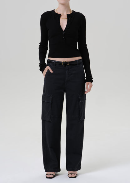 A model wearing a black long-sleeve cropped henley top and black wide-leg cargo pants with a black belt, standing against a grey background.