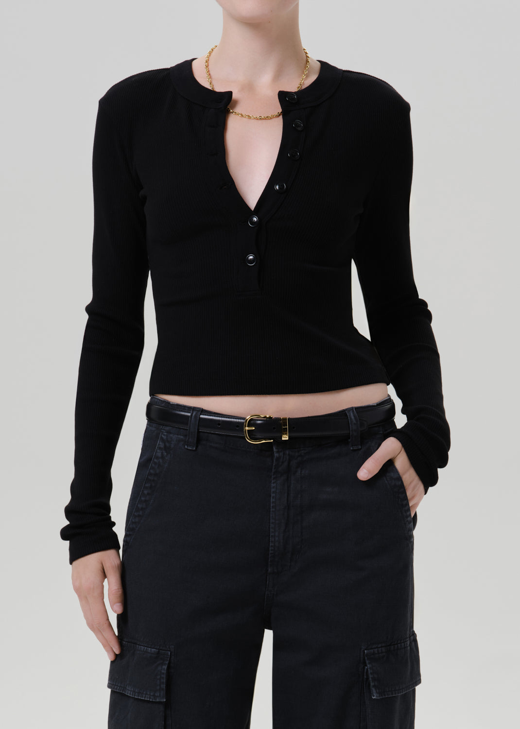 A model wearing a black long-sleeve crop top with a buttoned henley-style neckline, paired with black cargo pants and a black belt, against a grey background.