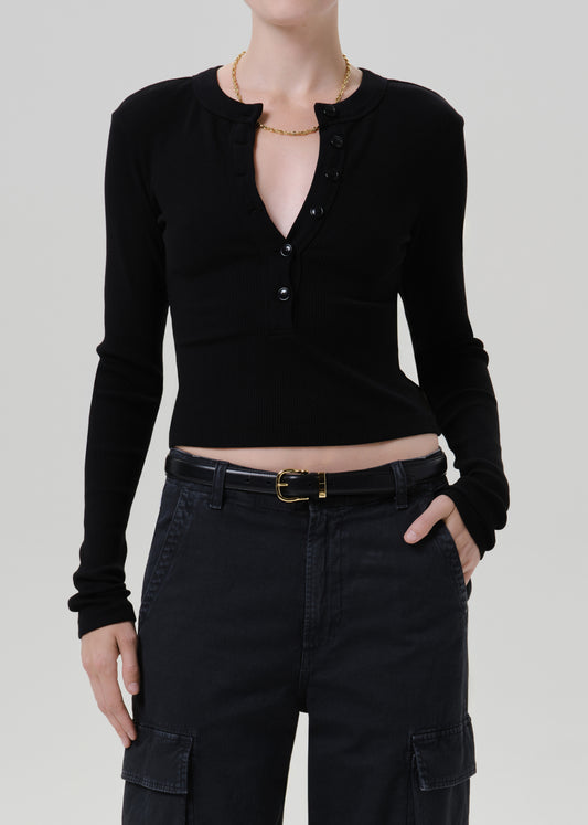 A model wearing a black long-sleeve crop top with a buttoned henley-style neckline, paired with black cargo pants and a black belt, against a grey background.