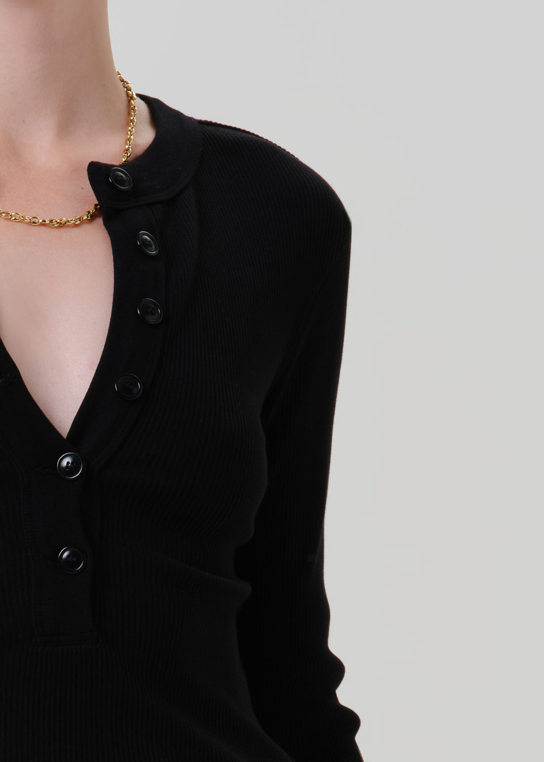 Close-up of a person wearing a black ribbed long-sleeve cardigan with a partial button placket and collar, paired with a gold chain necklace, against a grey background.