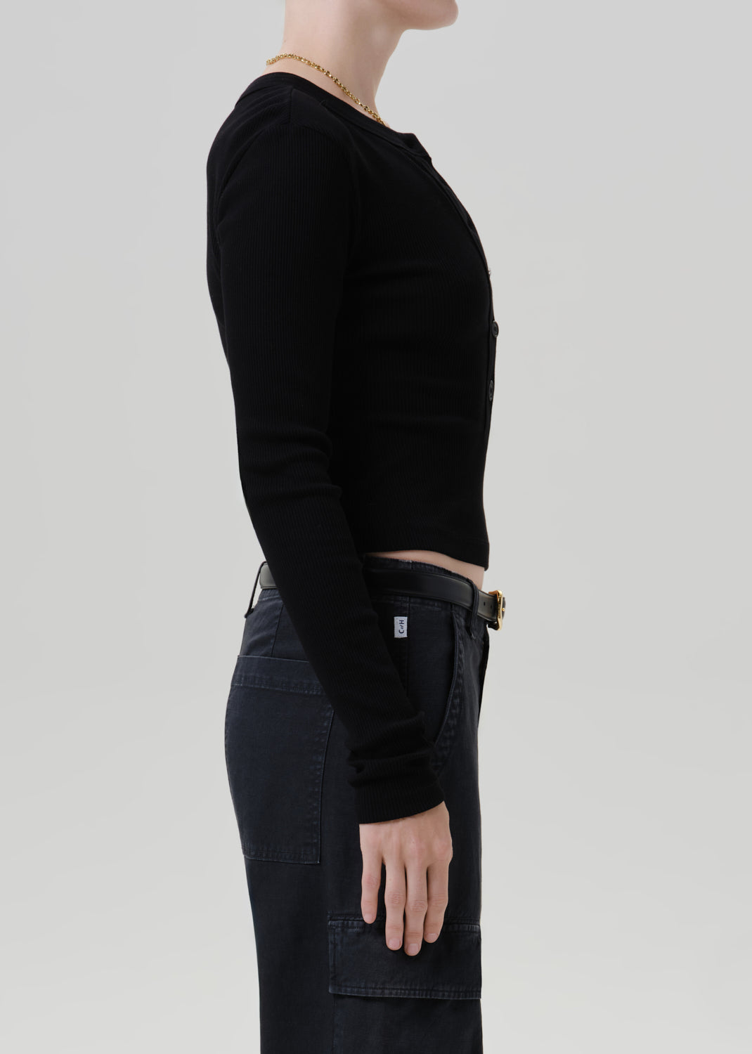 Side profile of a model wearing a black long-sleeve cropped top and dark denim cargo pants against a grey background.
