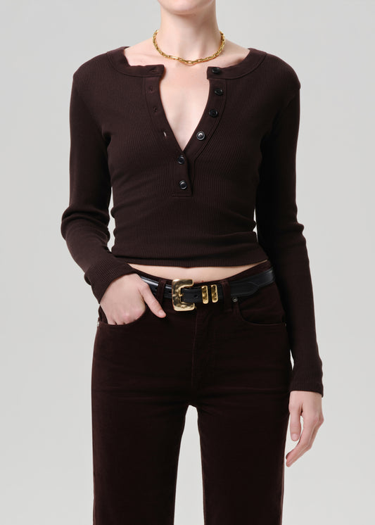 A model wearing a dark brown ribbed long-sleeve henley top with a split neckline and button placket, paired with dark brown pants and a black belt with a gold buckle, against a grey background.