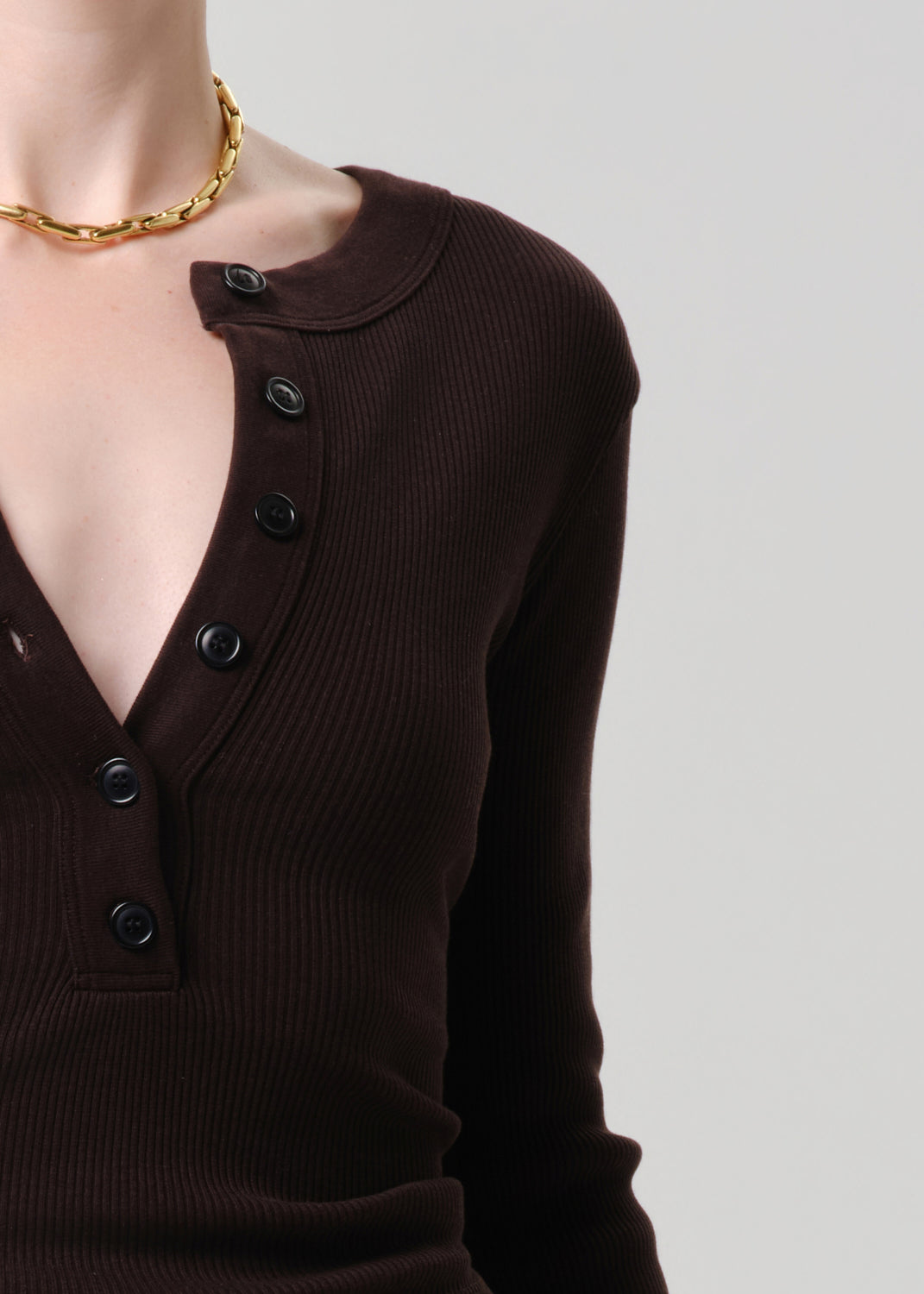 Close-up of a person wearing a dark brown ribbed long-sleeve henley top with black buttons, paired with a chunky gold chain necklace, against a grey background.
