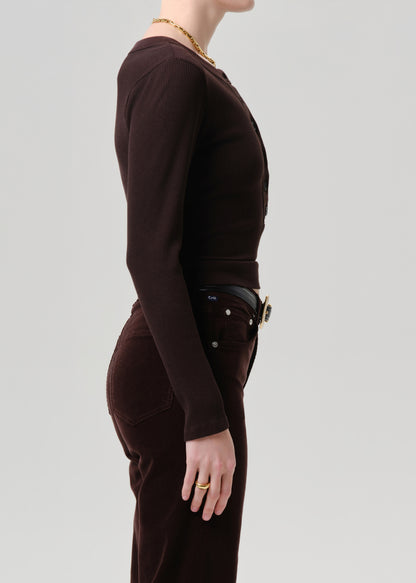 Side profile of a model wearing a dark brown ribbed knit long-sleeve top and matching dark brown trousers, accessorized with a gold chain necklace and rings, against a grey background.