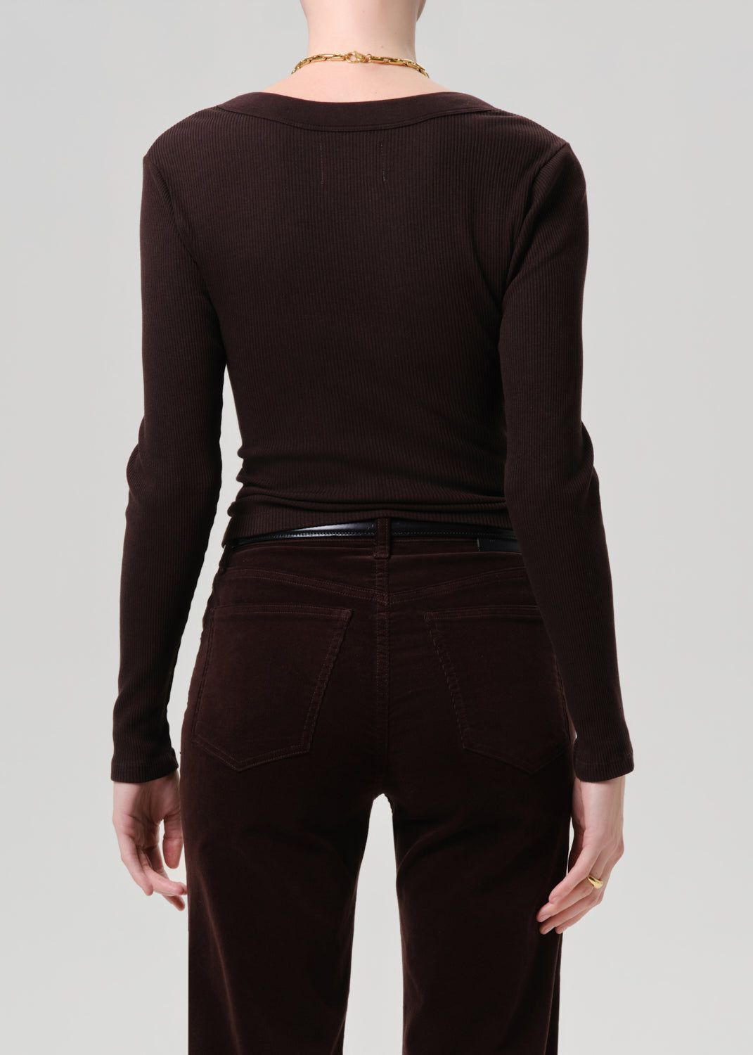 Rear view of a model wearing a dark brown long-sleeve knit top and matching dark brown corduroy pants against a grey background.