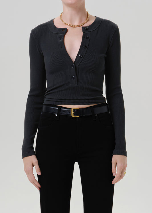 Person wearing a black long sleeve fitted button-front top with black pants and a black belt with gold buckle, front cropped view on a grey background.