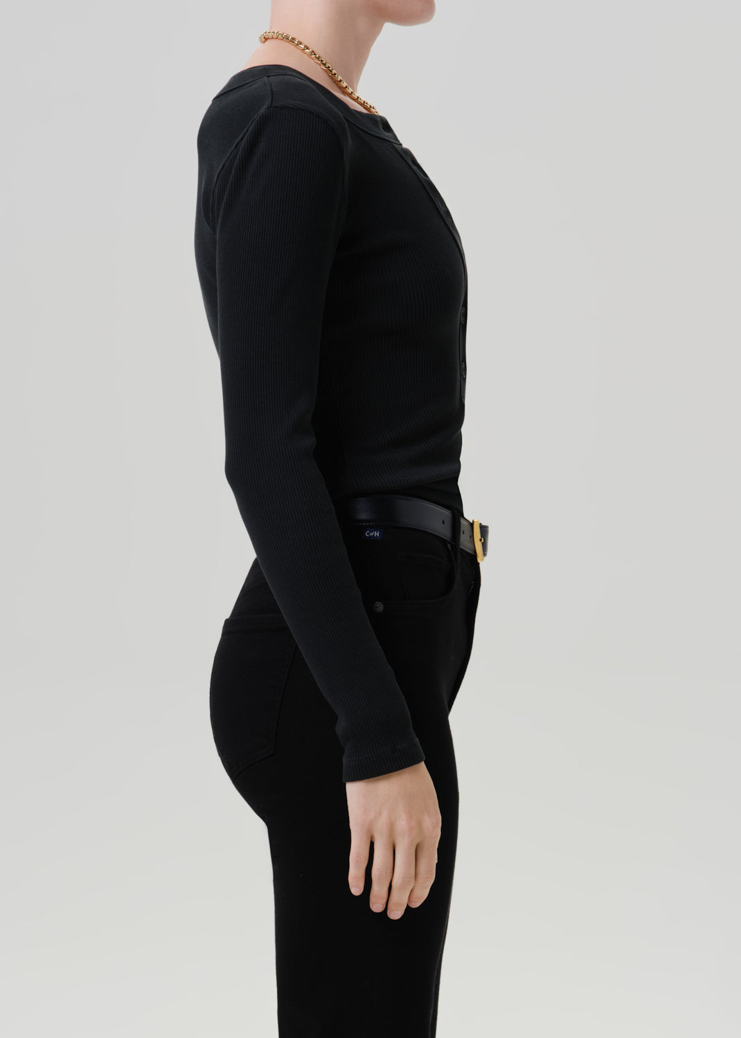 Person wearing a black long sleeve henley top with black pants viewed from the side on a grey background