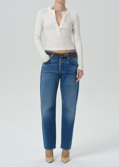 A model wearing a white long-sleeve henley top, blue straight-leg jeans with a brown belt, and beige heels, photographed against a grey background.