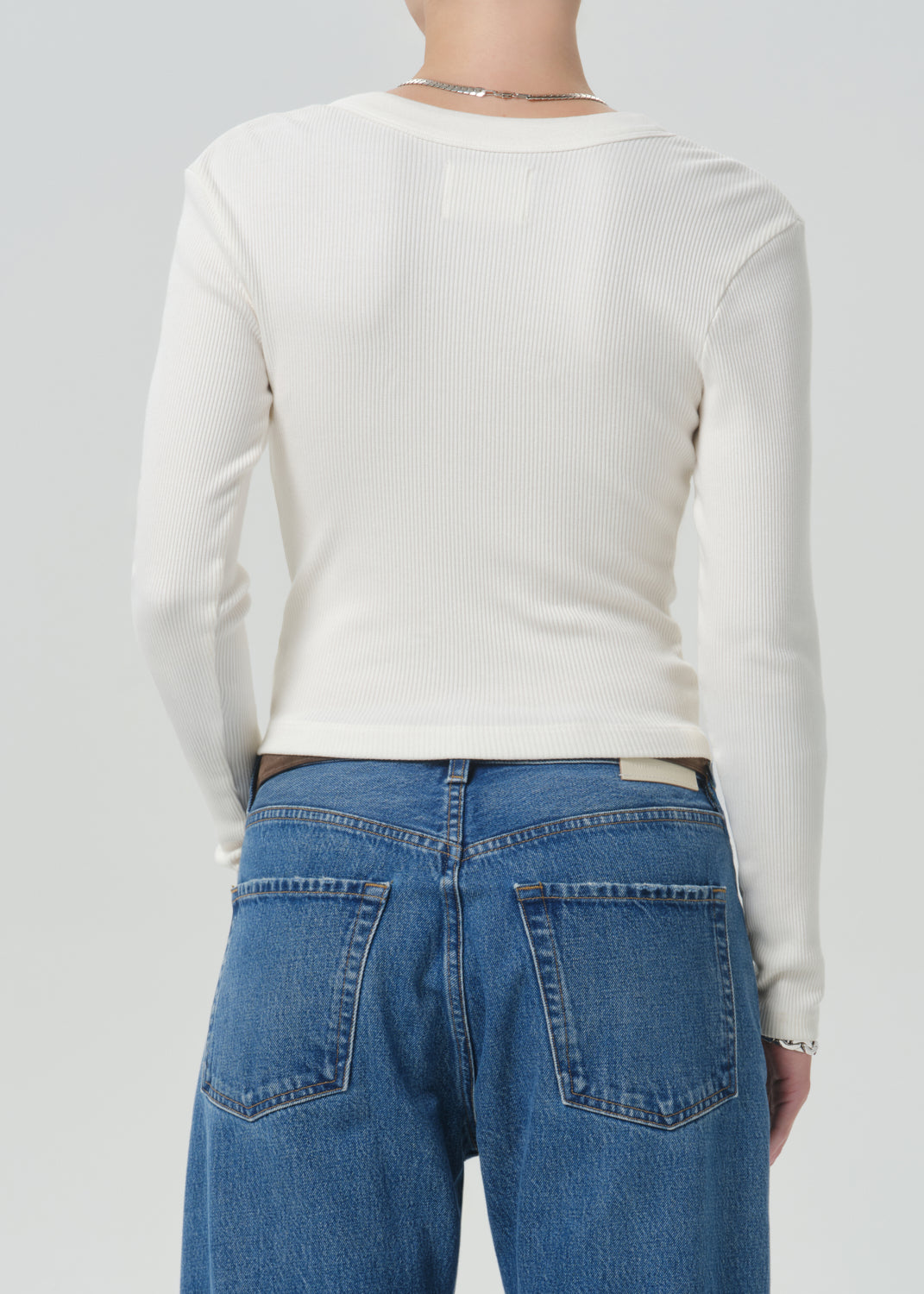 A person shown from the back wearing a fitted off-white ribbed long-sleeve top and blue jeans against a grey background.