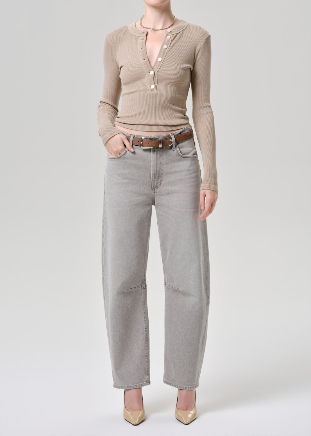 A model wearing a beige long-sleeve knit top with a split neckline and pearl-like buttons, paired with light grey wide-leg jeans and beige heels, shot against a light grey studio background.