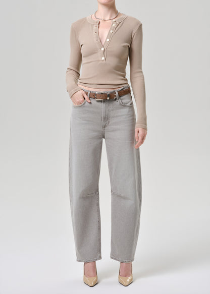 A model wearing a beige long-sleeve knit top with a split neckline and pearl-like buttons, paired with light grey wide-leg jeans and beige heels, shot against a light grey studio background.