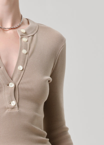 Close-up of a person wearing a ribbed knit long-sleeve henley top in a warm taupe-beige color, with white buttons down the front placket, against a light grey studio background.