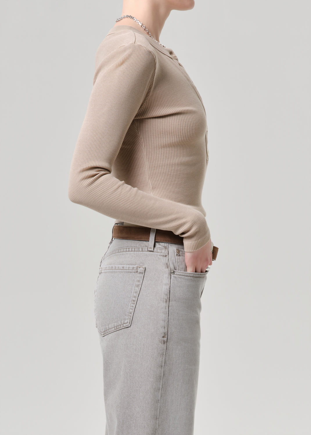 A side-profile view of a model wearing a beige ribbed long-sleeve top tucked into light grey jeans with a brown belt, against a solid light grey background.