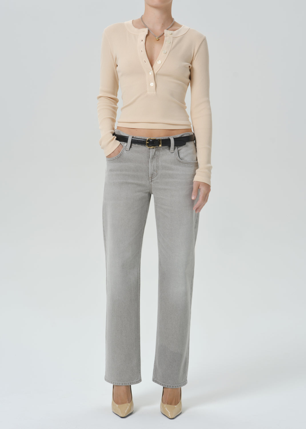 A model wearing a beige long-sleeve henley top and light gray straight-leg jeans with a black belt, paired with beige pointed-toe heels, against a grey background.