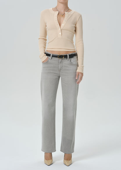 A model wearing a beige long-sleeve henley top and light gray straight-leg jeans with a black belt, paired with beige pointed-toe heels, against a grey background.