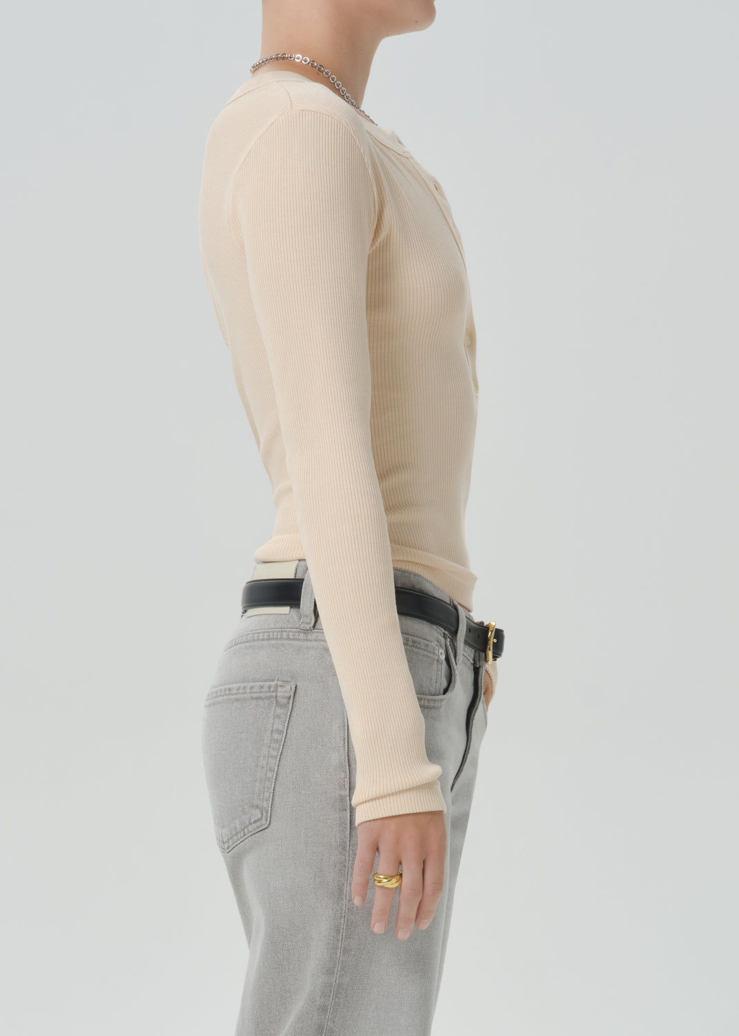 Side profile of a model wearing a beige long-sleeve ribbed knit top tucked into grey jeans with a black belt.