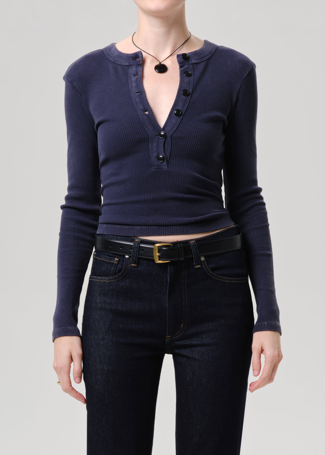 A model wearing a navy blue ribbed long-sleeve henley top with black buttons, paired with dark wash jeans and a black belt, against a grey background.