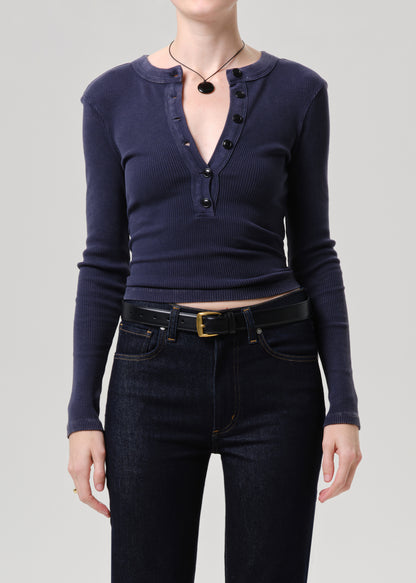 A model wearing a navy blue ribbed long-sleeve henley top with black buttons, paired with dark wash jeans and a black belt, against a grey background.