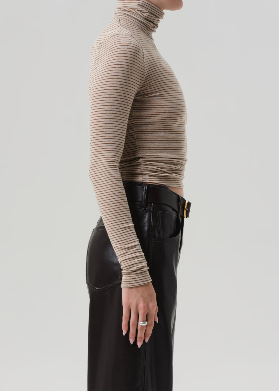Person wearing a beige ribbed turtleneck sweater and brown leather pants on a light gray background