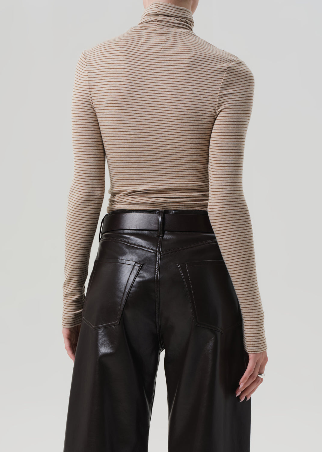 Person wearing a beige ribbed turtleneck sweater and brown leather pants on a light gray background