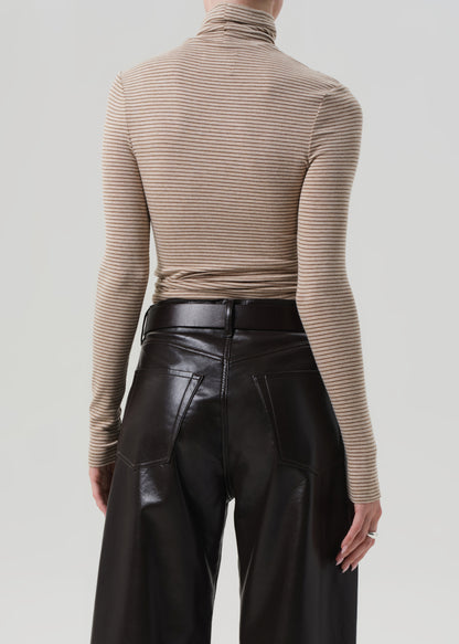 Person wearing a beige ribbed turtleneck sweater and brown leather pants on a light gray background