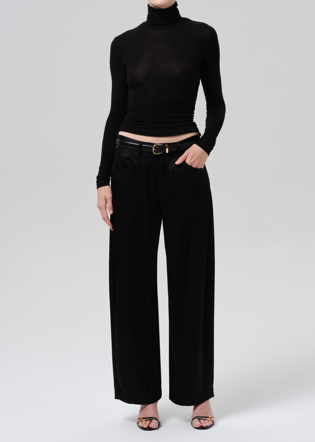 Person wearing a black fitted turtleneck with black wide leg trousers on a grey background