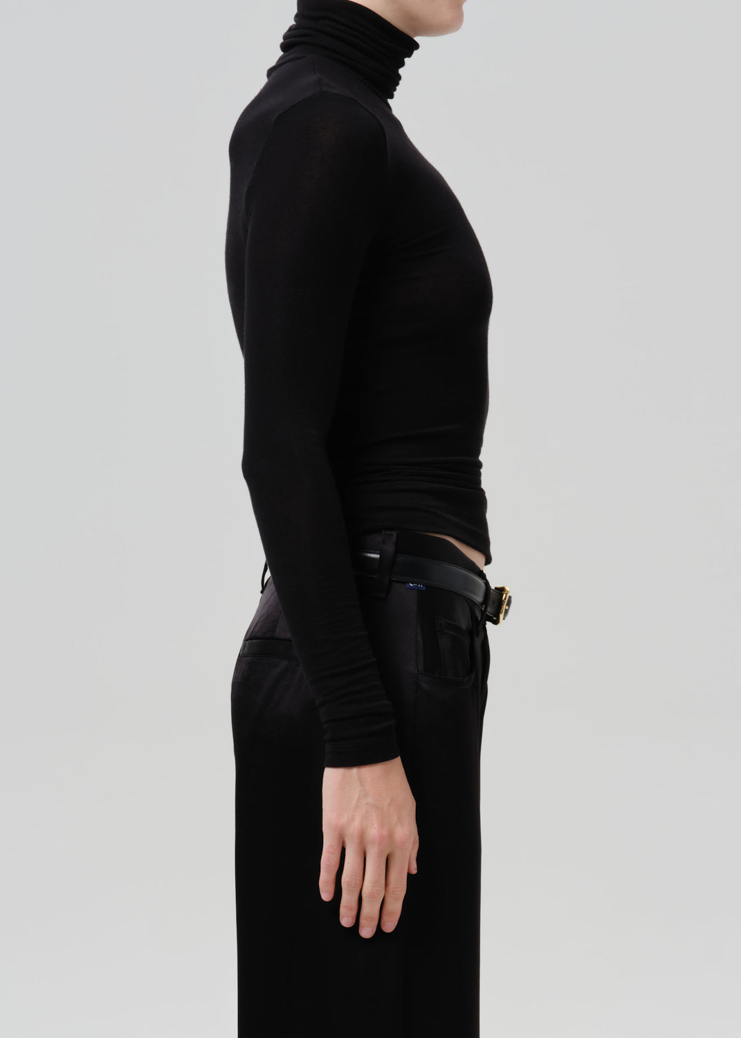 Person wearing a black fitted turtleneck with black wide leg trousers viewed from the side on a grey background
