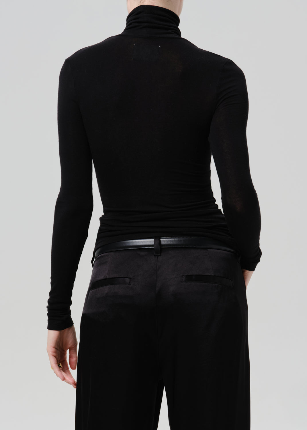 Person wearing a black fitted turtleneck with black wide leg trousers viewed from the back on a grey background
