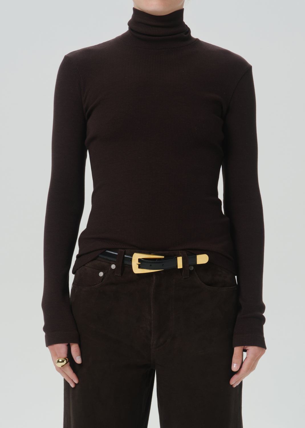 A person wearing a dark brown ribbed turtleneck sweater, dark brown pants, and a black belt with a gold buckle, shown from the neck down against a grey background.